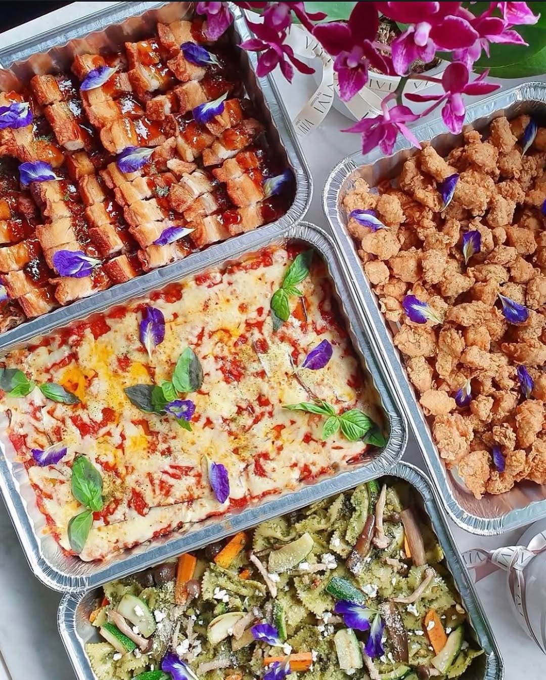 Flossom catering spread — lechon, pasta, and sides with butterfly pea garnish